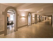 Langer Flur mit gewölbeartigen Decken und beleuchteten Wandnischen, die durch Rundbögen unterteilt sind; links Einblicke in einen Raum mit Tisch und Stühlen, rechts und hinten eine klare Sicht auf die glatten Wände und den polierten Boden.