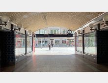 Ein überdachter Durchgang mit gelbem Klinkermosaik an der Decke, flankiert von Glasfenstern und Wandleuchten mit runden Lampenschirmen. Am Ende des Durchgangs sind Straßenszenen mit Fußgängern sowie ein modernes Gebäude mit großen Fenstern sichtbar.