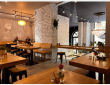 Ein modernes Café mit hellen Holzmöbeln, weißen Backsteinwänden und großem Kronleuchter. Im Hintergrund sind Sitzbänke, dekorative Pflanzen und große Fenster mit gewölbten Rahmen zu sehen.