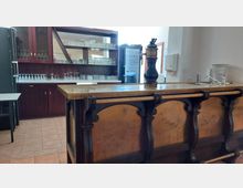 Barbereich mit einer Theke aus Holz und Metallstreben, darauf eine Bierzapfanlage. Im Hintergrund befinden sich Holzregale mit Gläsern sowie ein Kühlschrank, flankiert von weißen Wänden und Fliesenboden.