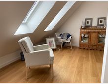 Zimmer mit Dachschrägen und zwei großen Dachflächenfenstern, ausgestattet mit einem hellen Polstersessel, einem kleinen Schränkchen aus Holz, einem Wäschekorb und einem Holzfußboden. An der Wand steht ein gerahmtes Bild auf dem Boden.