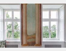 Innenraumansicht mit zwei großen weißen Sprossenfenstern, die eine grüne Landschaft zeigen, und einer Wand dazwischen, die mit einer dekorativen Malerei antiker Säulen gestaltet ist. Unter den Fenstern befinden sich Heizkörper mit Zierverkleidungen.