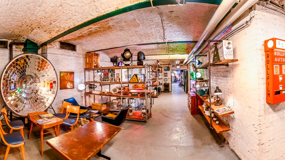 Gewölbekeller mit weiß gestrichenen Ziegelwänden und sichtbaren Rohrleitungen an der Decke, eingerichtet mit Vintage-Möbeln, Tischen und Stühlen sowie Regalen voller Industrieleuchten und Objekte. Links steht eine große runde Spiegel- oder Reflektorscheibe, rechts hängt ein roter „Rollfilm-Automat“ an der Wand; ein langer Gang führt in weitere Räume.