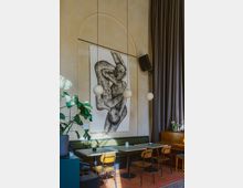 Ein gemütliches Café mit einer Sitzbank, Holztischen und Stühlen vor einer beigefarbenen Wand, geschmückt mit einem großen schwarz-weißen Kunstwerk. Drei runde Hängelampen hängen von der Decke, und Pflanzendekoration sowie ein Klavier befinden sich im Hintergrund.