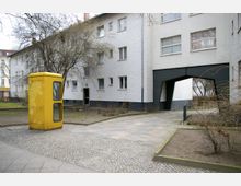 Gelbe Telefonzelle auf einem gepflasterten Platz vor einem mehrstöckigen Wohngebäude mit weißer Fassade und einem Durchgangsbogen. Im Hintergrund befinden sich kahler Baumbewuchs und ein kleiner Hofbereich.