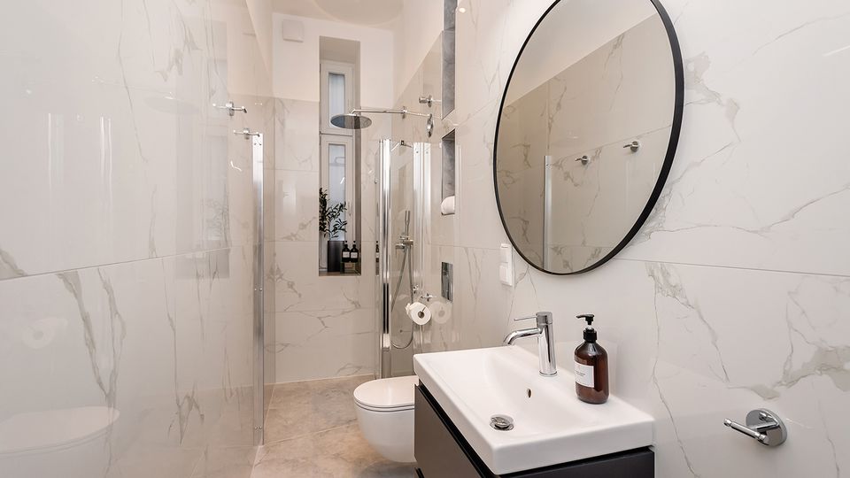Schmales, modernes Badezimmer mit großformatigen, marmorierten Wandfliesen, bodengleicher Glasdusche mit Regendusche und einem Waschbecken auf dunklem Unterschrank. Ein runder Wandspiegel, ein wandhängendes WC und ein hohes Fenster am Ende des Raums ergänzen die helle, minimalistische Ausstattung.