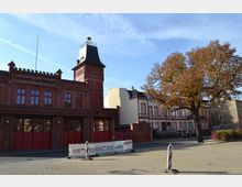 Backsteingebäude eines Feuerwehr-Gerätehauses mit rotem Tor und einem Turm, daneben historisch wirkende Wohnhäuser mit verzierten Fassaden und ein großer Baum mit Herbstlaub im Vordergrund. Der Himmel ist klar und blau.