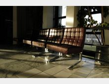 Reihe von braunen Ledersesseln mit Metallrahmen in einer modernen Wartehalle; im Hintergrund große Fenster, durch die Tageslicht fällt, sowie Pflanzen und ein Blick auf eine Wiese und Gebäude draußen. Fliesenboden im Vordergrund.