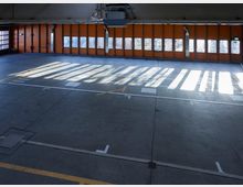 Große, leere Halle mit Betonboden und einer gegenüberliegenden Wand aus orangefarbenen Toren mit Glasfenstern. Lichtstrahlen von außen werfen Schatten von den Fensterelementen auf den Boden, und Markierungen sind auf dem Boden sichtbar.