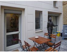 Ein kleiner Innenhof mit hellem Gebäude im Hintergrund, das zwei Fenster mit Gitter und eine Glastür zeigt. In der Mitte steht ein Holztisch mit vier Klappstühlen, daneben eine bronzene Statue einer stehenden Person.