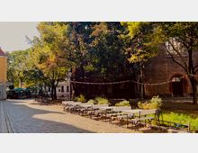 Kopfsteingepflasterter Platz mit Bäumen, Sitzgelegenheiten und einem Ziegelfassade-Gebäude im Hintergrund. Zwischen den Bäumen hängen bunte Wimpel, und links sind ein Cafébereich mit Sonnenschirmen und Fahrräder zu sehen.