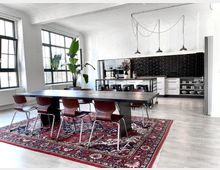 Großzügiger, gut beleuchteter Raum im Industriestil mit weißen Wänden, großen Fenstern und einer offenen Küche mit schwarzen Fliesen. Im Vordergrund ein langer Esstisch mit Holzstühlen auf einem roten Perserteppich, daneben steht eine große Grünpflanze.