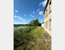 Ebene Wiese mit einem schmalen Weg neben einem Gebäude mit vergitterten Fenstern, im Hintergrund ein Fluss sowie Büsche und Bäume unter klarem, blauem Himmel.