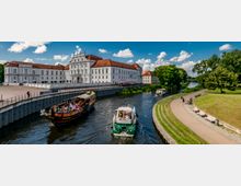 Historisches Schlossgebäude mit weißer Fassade und roten Dächern an einem Fluss. Im Vordergrund fahren Boote auf dem Wasser, während rechts ein gepflegter Park mit Bänken und Spazierwegen zu sehen ist.