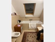 Ein Badezimmer mit beige gefliesten Wänden und einer Badewanne unter einem Dachfenster aus Holz. Im Vordergrund stehen eine Toilette, eine kleine Holzkommode mit lila Toilettenpapier darauf und ein gemusterter Teppich auf den Fliesen.