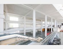 Weitläufiges, modernes Gebäude mit weißem Interieur, offenen Galerien und großen Glasfenstern, die viel Tageslicht hereinlassen. Im Vordergrund sind Treppen mit Metallgeländern und ein dunkler Steinboden sichtbar.