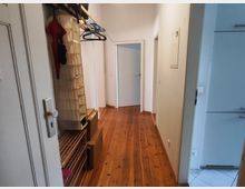 Narrow apartment hallway with white walls and a varnished wooden plank floor, leading to a closed door at the end. On the left are open coat hooks and a shoe rack, with doorways opening off the corridor on the right.
