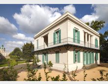 Klassizistisches Gebäude mit weißer Fassade, grünen Fensterläden und umlaufendem Balkon, umgeben von einem Garten mit Wegen und Pflanzen; im Hintergrund ist der Kuppelturm eines weiteren historischen Gebäudes zu sehen.