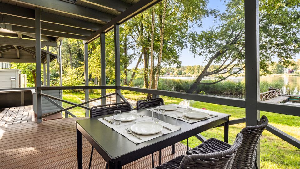 Überdachte Holzterrasse mit Tisch und geflochtenen Stühlen, gedeckt mit Tellern und Gläsern. Blick durch das Geländer auf eine grüne Uferlandschaft mit Bäumen und Schilf am See sowie einen kleinen Steg am Wasser.