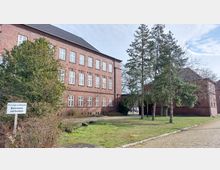 Backsteingebäude mit mehreren weißen Fenstern und dunkelgrauem Dach umgeben von Bäumen und Grünfläche; im Vordergrund ein Schild mit der Aufschrift "Privatgrundstück Betreten verboten".