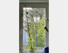 Ein Blick aus einem Fenster mit weißem Rahmen, im Vordergrund eine Zimmerpflanze in einem Topf und Glasgegenstände. Draußen sichtbar ist eine ältere schwarze Laterne vor grünem Gebüsch und Bäumen, sowie ein Teil eines Hauses mit blauem Anstrich und rotem Dach.