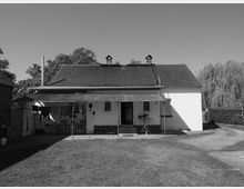 Schwarzweißfoto eines einfachen Einfamilienhauses mit Satteldach und weißer Fassade. Vor dem Haus befindet sich ein überdachter Sitzbereich mit Wäschestücken und Gartenmöbeln; im Hintergrund sind Bäume und ein gepflegter Rasen erkennbar.