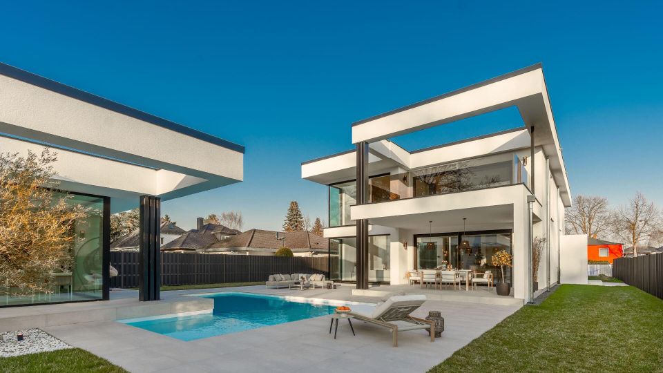 Moderner, zweigeschossiger Flachdachbau mit großen Glasfronten und umlaufendem Balkon, der sich zu einer gepflasterten Terrasse mit rechteckigem Swimmingpool öffnet. Der Außenbereich ist von Rasenflächen und einem hohen Zaun umgeben, im Hintergrund sind Nachbarhäuser und Bäume zu sehen.