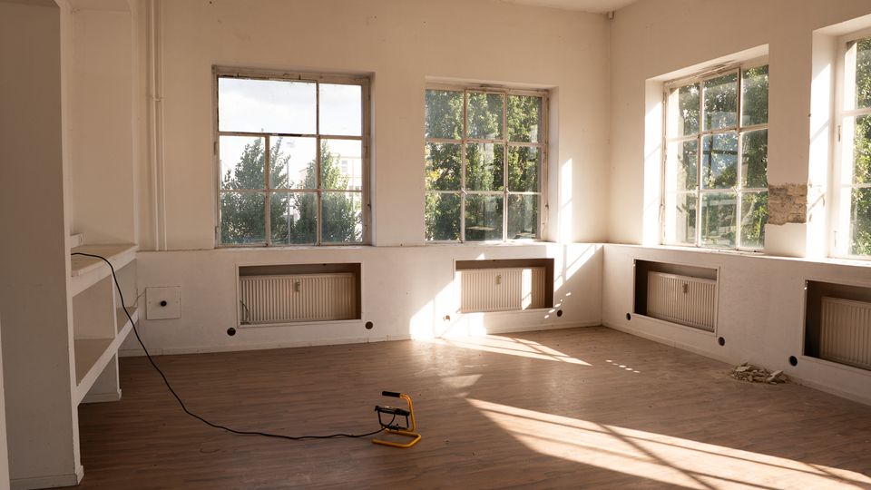 Heller, weitgehend leerer Innenraum mit Holzboden, weißen Wänden und mehreren großen Sprossenfenstern, durch die Bäume zu sehen sind. Unter den Fenstern sind eingelassene Heizkörper, und Sonnenlicht wirft lange Schatten in den Raum.