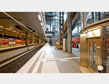 Modernes Bahnsteigambiente in einem überdachten Bahnhof mit mehreren Gleisen, einem roten Regionalzug auf der rechten Seite und einem gläsernen Aufzug im Hintergrund. Das Design umfasst hohe Säulen, glatte Metallverkleidungen und Sitzbänke entlang des Bahnsteigs.