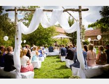 Eine Hochzeit im Freien mit Gästen, die auf weißen Bänken sitzen, und einem dekorierten Birkenbogen mit weißem Stoff im Vordergrund. Im Hintergrund befinden sich ein Redner, ein Sonnenschirm, Bäume und ein Ziegelgebäude mit roten Dächern.