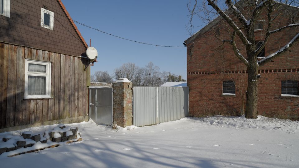 Verschneiter Hof zwischen einem Holzhaus mit Satteldach und einem Backsteingebäude, getrennt durch ein hohes Metalltor und einen gemauerten Torpfeiler. Im Vordergrund liegt Schnee, rechts steht ein kahler Baum vor der Backsteinfassade unter klarem Himmel.