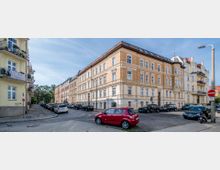 Straßenszene mit mehrstöckigen Wohnhäusern im Altbaustil, geprägt von hellen Fassaden mit Backsteinelementen und zahlreichen Fenstern. Geparkte Autos säumen die Straßenränder, und im Hintergrund ist vereinzelt Grün zu erkennen.