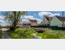 Kleinstädtische Wohngegend mit Einfamilienhäusern, Gärten und einer gepflasterten Brücke über einen kleinen Bewässerungsgraben. Im Vordergrund sind ein Feldweg, eine Wiese und ein Baum zu sehen, im Hintergrund helle Häuser mit roten und braunen Ziegeldächern.