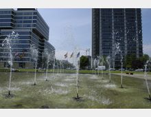 Moderner Universitätscampus mit einem rechteckigen Wasserbecken im Vordergrund, aus dem mehrere kleine Fontänen sprudeln. Im Hintergrund befinden sich zwei mehrstöckige Gebäude mit Glasfassaden, eines davon mit der Aufschrift „Universität“, sowie Flaggen und Grünflächen.