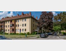 Mehrfamilienhaus mit gelber Fassade und rotem Satteldach in einer Wohnstraße mit parkenden Autos und Bäumen. Rechts fährt ein Fahrradfahrer auf der asphaltierten Straße entlang.