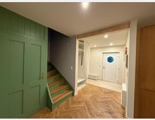 Wohnraum mit modernem Eingangsbereich, einer weißen Tür mit rundem Fenster, Holzfußboden in Fischgrätmuster, einer grünen Holzverkleidung an der Wand und einer Treppe mit Holzstufen, die nach oben führt. Ein offener Einbauschrank mit Vorhang befindet sich neben der Tür.