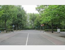 Kreuzung in einer parkähnlichen Umgebung mit Gehwegen, Ampeln und Straßenschildern, umgeben von zahlreichen Bäumen und Grünflächen.