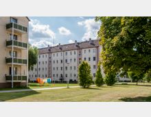 Mehrstöckige Wohngebäude mit hellen Fassaden und grauen Dächern, umgeben von einer Grünfläche mit Bäumen und Wäscheleinen. Im Vordergrund sind ein gepflasterter Weg und ein großer Baum zu sehen.