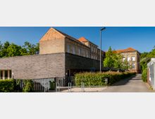 Backsteinbau mit einem modernen Anbau aus dunklen Ziegeln, beschriftet mit „Haus der kleinen Forscher“. Der historische Teil des Gebäudes hat hohe Fenster und ein rotes Ziegeldach. Im Vordergrund befinden sich ein Gehweg, eine Hecke und ein Zaun, umgeben von Bäumen bei sonnigem Wetter.
