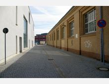 Schmale Gasse mit Kopfsteinpflaster, flankiert von einem modernen weißen Gebäude mit großen Glasfenstern auf der linken Seite und einem gelben Backsteingebäude mit klassischen Fenstern auf der rechten Seite; im Hintergrund sind moderne, rot-schwarze Gebäude zu sehen.