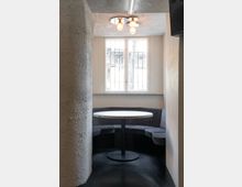 Kleine, minimalistische Sitzecke mit einem runden Tisch aus hellem Material und einer halbrunden, gepolsterten Sitzbank. Im Hintergrund befindet sich ein vergittertes Fenster, darüber eine moderne Deckenleuchte mit drei Glühbirnen.