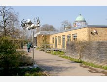 Backsteinbau mit großen Fenstern und Rampe, umgeben von einer gepflasterten Gehwegfläche und spärlicher Vegetation. Im Hintergrund ist eine grüne Kuppel eines historischen Gebäudes sowie eine moderne Metallskulptur auf einem Ständer zu sehen.