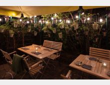 Beleuchtete Terrasse mit Holztischen und -stühlen, dekoriert mit Kerzenlampen und Geschirr. An einem Metallgitter im Hintergrund hängen Blumentöpfe mit Kräutern und Pflanzen, umgeben von Lichterketten unter einer gelben Markise.