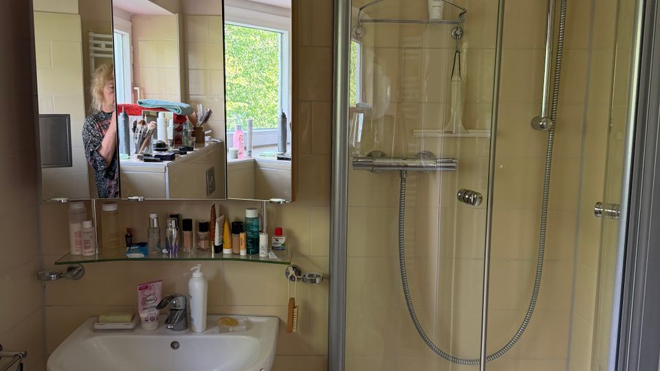 Kleines Badezimmer mit beigefliesten Wänden, Waschbecken mit Spiegel- und Glasablage voller Pflegeprodukte sowie einer Eckdusche mit gebogener Glasabtrennung. Ein Fenster sorgt für Tageslicht und spiegelt sich im Spiegelschrank.