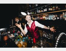 Innenansicht einer dunklen Cocktailbar mit schwarzer Fliesenwand, langen Regalreihen voller Spirituosenflaschen und warmen Lichtleisten. Im Vordergrund steht eine dicht bestückte Barstation mit Flaschen, Shakern, Eis und Barwerkzeugen.