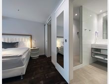 Modernes Hotelzimmer mit einem Bett auf der linken Seite, einem Nachttisch mit Lampe und einem angrenzenden Badezimmer. Das Badezimmer verfügt über eine Glasdusche, einen Waschtisch mit Spiegel und helle Fliesen.