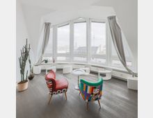 Heller moderner Raum mit großen, halbkreisförmigen Fenstern, die einen städtischen Ausblick bieten, sowie zwei bunten Designerstühlen, einem kleinen Tisch und einem großen Kaktus in einem Topf. Im Hintergrund sind weiße Sitzmöbel entlang der Fensterfront angeordnet.