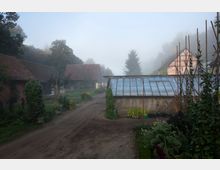 Ein ländlicher Hof mit mehreren Gebäuden, darunter ein Gewächshaus mit Glasdach rechts und Ziegeldachhäuser im Hintergrund. Der Boden besteht aus einem unbefestigten Weg, umgeben von Pflanzen, Gemüsebeeten und dichtem Laub, bei leichtem Nebel in ruhiger Landschaft.