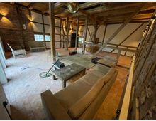 Rustikaler Innenraum eines Fachwerkhauses mit freiliegenden Holzbalken, Ziegelwänden und einem Ziegelboden. Der Raum ist mit verschiedenen Sitzmöbeln, darunter Sofas und Korbstühle, sowie einem Holztisch ausgestattet.