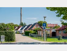 Wohnsiedlung mit kleinen Einfamilienhäusern, geordnet entlang einer gepflasterten Straße. Im Hintergrund ragt ein hoher Industrieschornstein auf, umgeben von Bäumen und einem klaren, blauen Himmel; Straßenschilder und Zäune markieren die Grundstücke.